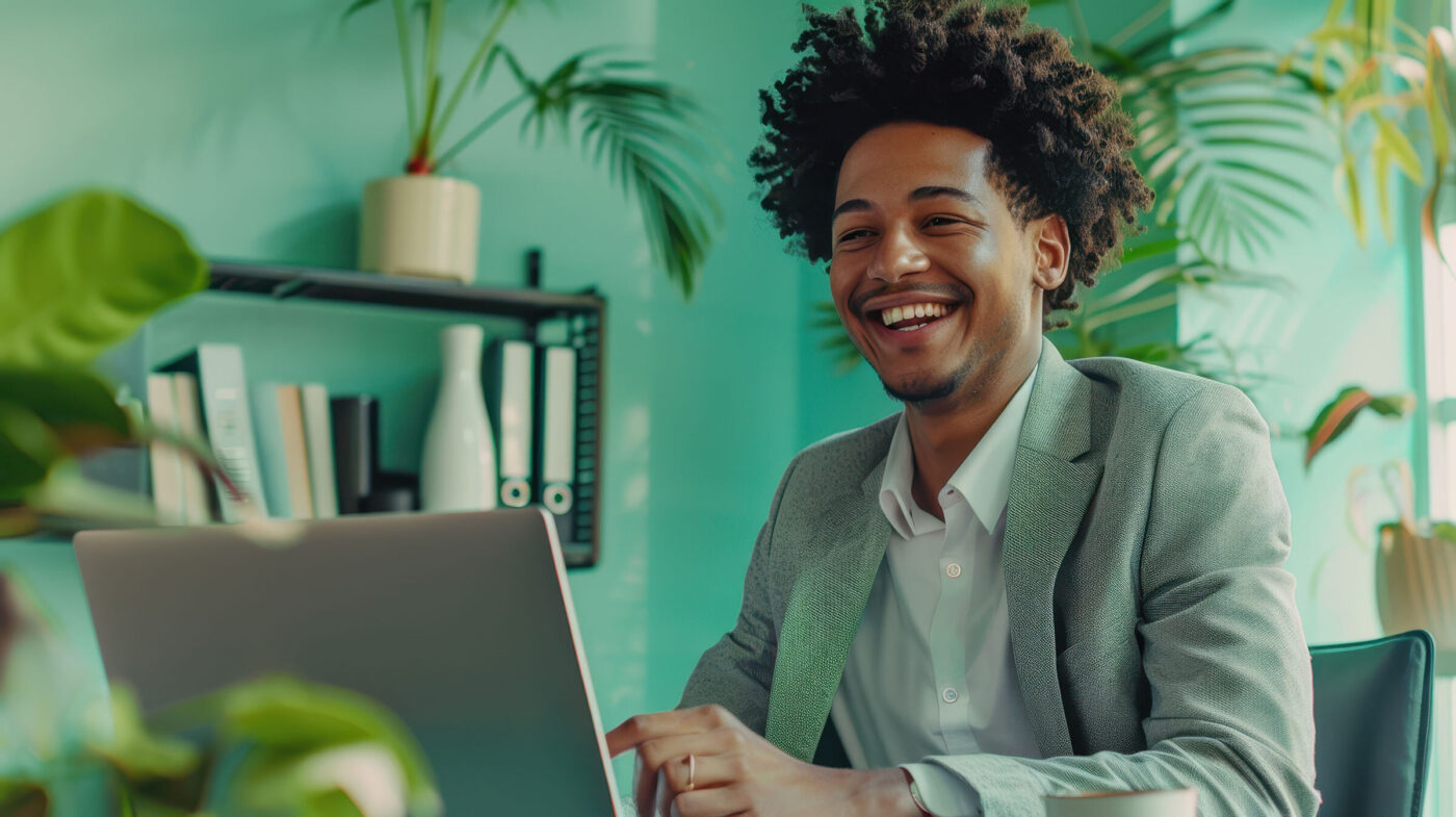Hombre joven con traje gris sonríe mientras trabaja en una laptop, rodeado de plantas en una oficina moderna
