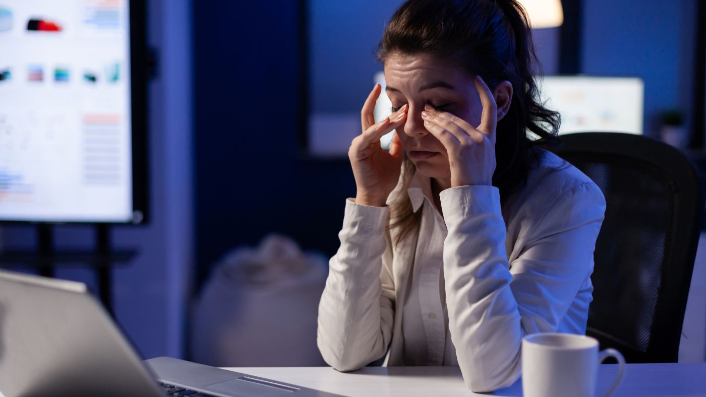 Mujer en oficina frente a laptop mostrando signos de cansancio, reflejando el impacto del estrés laboral en la salud y la preocupación de las empresas