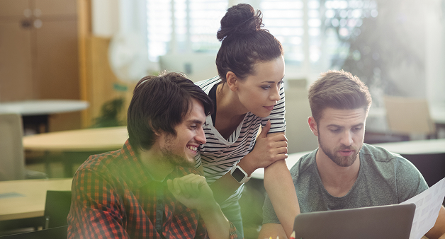Tres personas jóvenes reunidas alrededor de un computador portátil en un entorno de oficina iluminado. Uno de ellos sostiene una hoja mientras los otros observan atentamente la pantalla, colaborando en equipo con actitud concentrada y positiva.