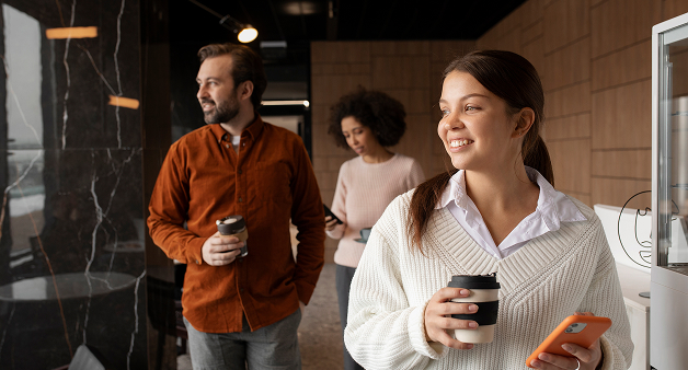Grupo diverso de personas en una oficina moderna. Una mujer en primer plano sonríe con café y celular en mano, transmitiendo bienestar laboral y cultura organizacional positiva.