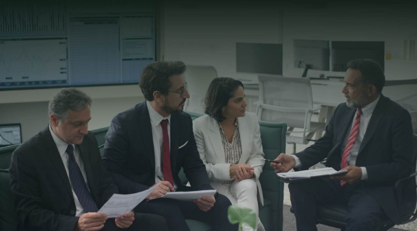 Reunión estratégica de equipo corporativo con cuatro profesionales diversos sentados en sofás verdes en una oficina moderna. Un hombre de negocios mayor a la derecha habla y sostiene una tableta. Los otros miembros escuchan. El fondo muestra un monitor grande con análisis de datos financieros y gráficos de velas. Imagen de fondo de banner corporativo profesional.