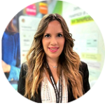Retrato profesional de perfil circular de una mujer con cabello rubio ondulado, vistiendo un blazer negro sobre una blusa blanca estampada, sonriendo a la cámara en un entorno de evento corporativo. Lleva una credencial (lanyard) colgando del cuello y de fondo se aprecian pantallas borrosas con gráficos comerciales